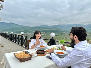 Una mesa de comedor con comida y bebidas, una barandilla y vistas a colinas verdes.