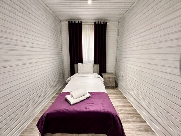 Habitación individual estrecha, con piso de madera y paredes blancas, que presenta una colcha morada y una toalla blanca sobre la cama.