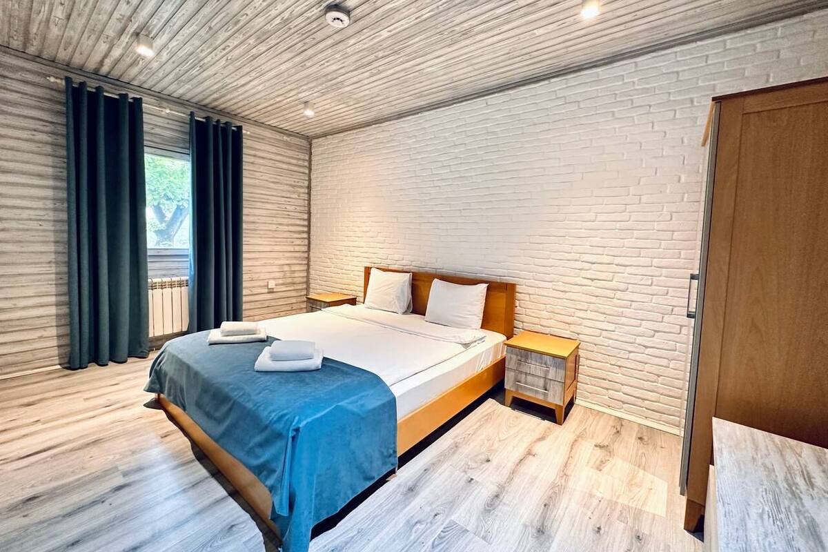 A modern bedroom with a wooden ceiling, a brick-patterned wall, a bed with a blue bedspread, and a wooden nightstand.