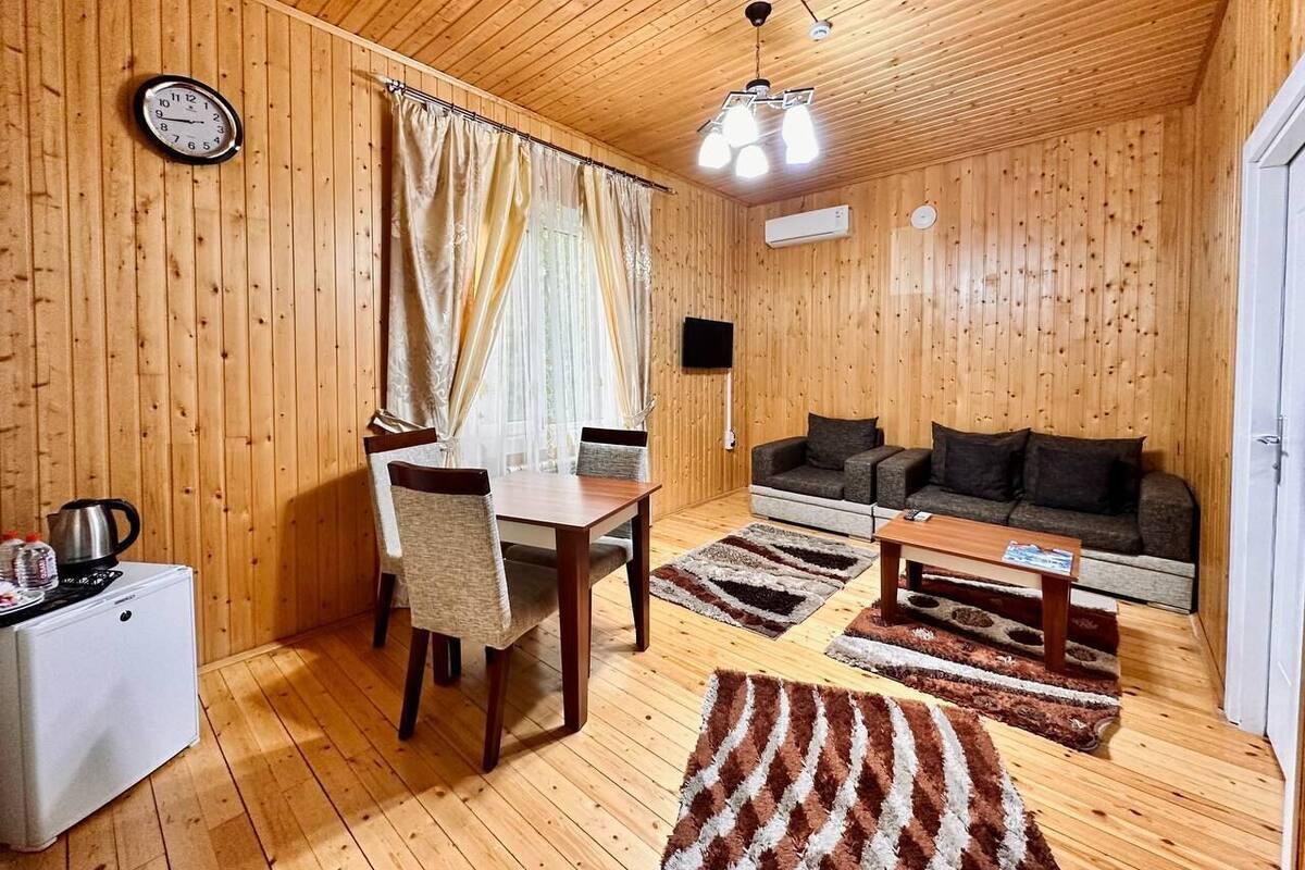 A cozy room with wooden walls and ceiling, a dining table with chairs, a sofa, and a small white fridge.