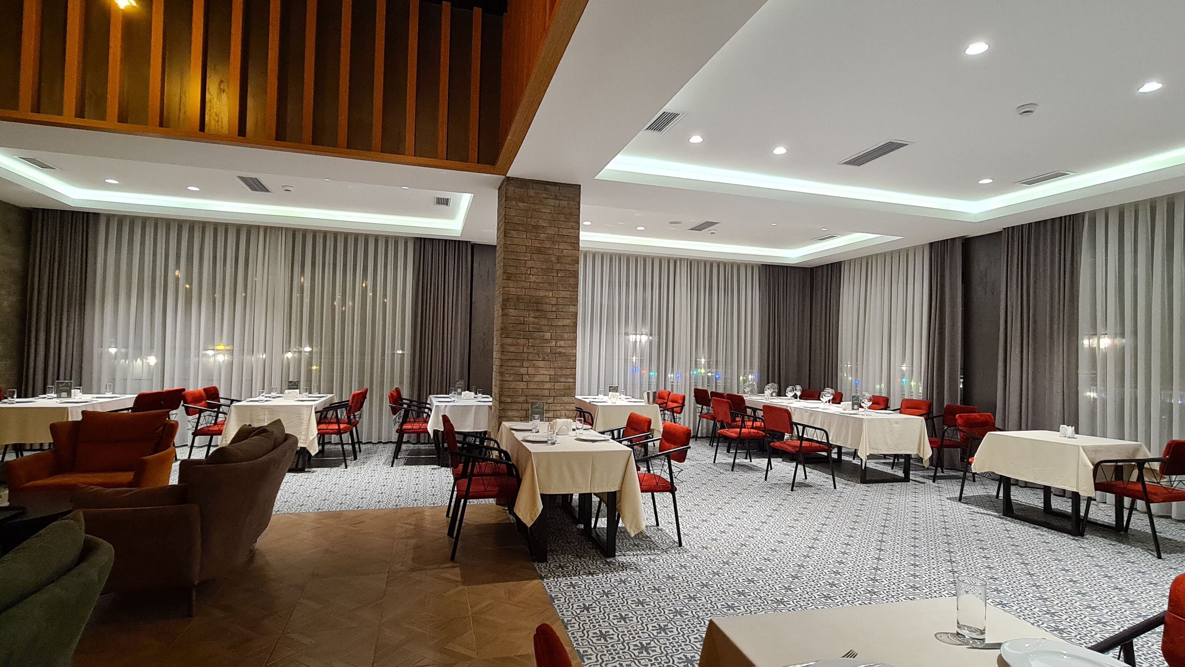 A hotel dining area with multiple tables and chairs, large windows with curtains, and a patterned floor.