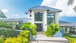 A modern two-story house with a flat roof, large windows, and a well-maintained garden with yellow flowering plants and palm trees. - Ocean Pearl 4 Bedroom Home by RedAwning (Roatan)