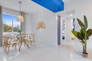 Un'area pranzo moderna con tavolo e sedie bianche, un lampadario a sospensione e un'ampia finestra con vista su edifici e vegetazione.