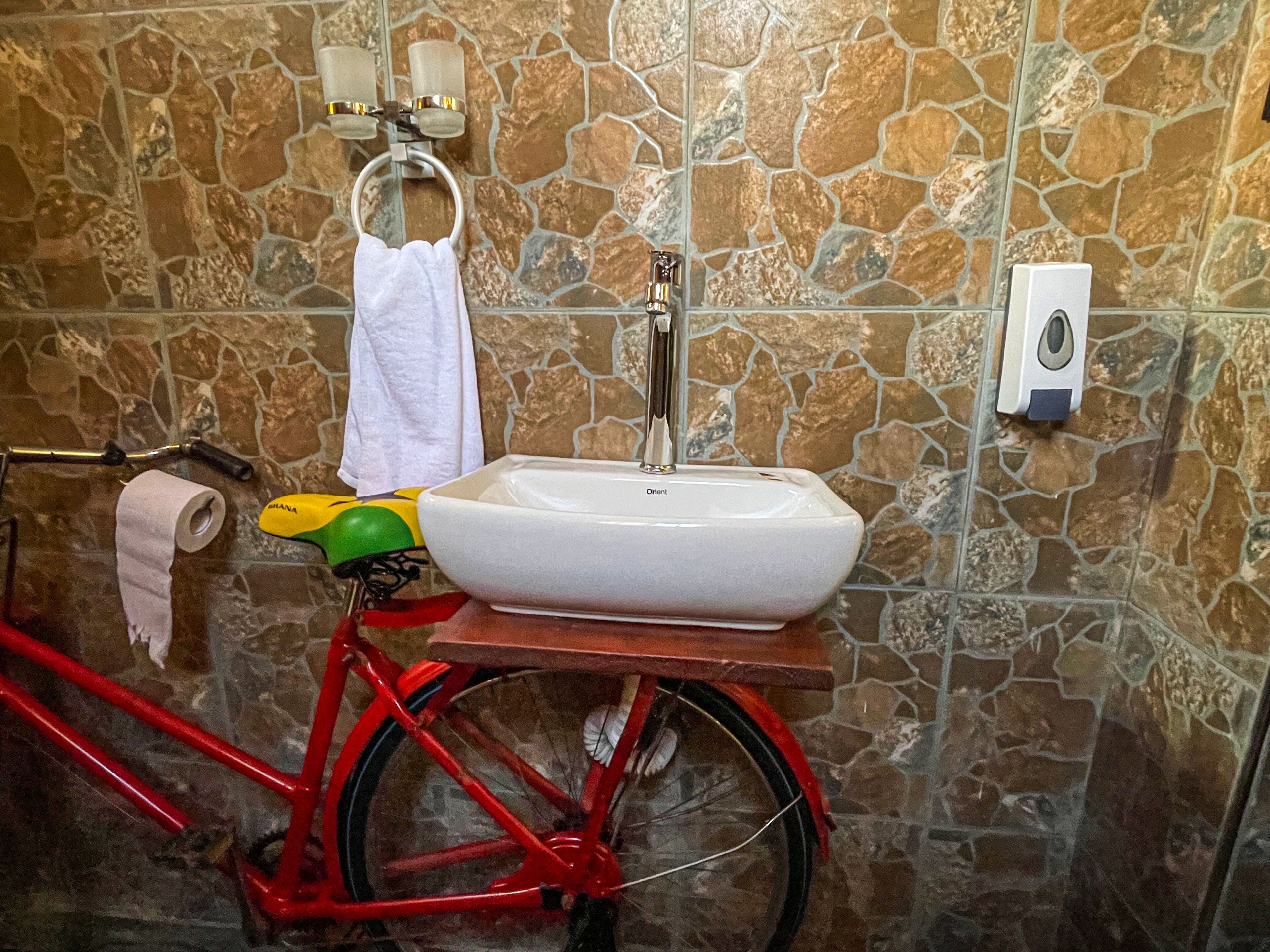 Une salle de bain avec un mur en pierre, un lavabo blanc, un vélo rouge, une serviette et un rouleau de papier toilette.
