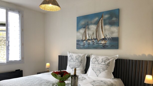 Een net opgemaakt bed met beddengoed met een patroon, een schilderij van zeilboten aan de muur en een vaas met bloemen op het nachtkastje.
