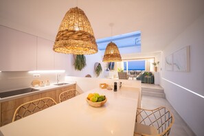 A modern kitchen with a central island, wicker pendant lights, and a view of the sea through a large window.