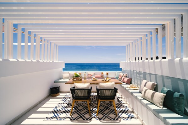 A sunlit outdoor dining area with a view of the sea, featuring a wooden table, two chairs, and a bench with cushions.