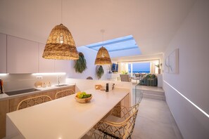 A modern kitchen with a central island, wicker pendant lights, and a view of the sea through a skylight.