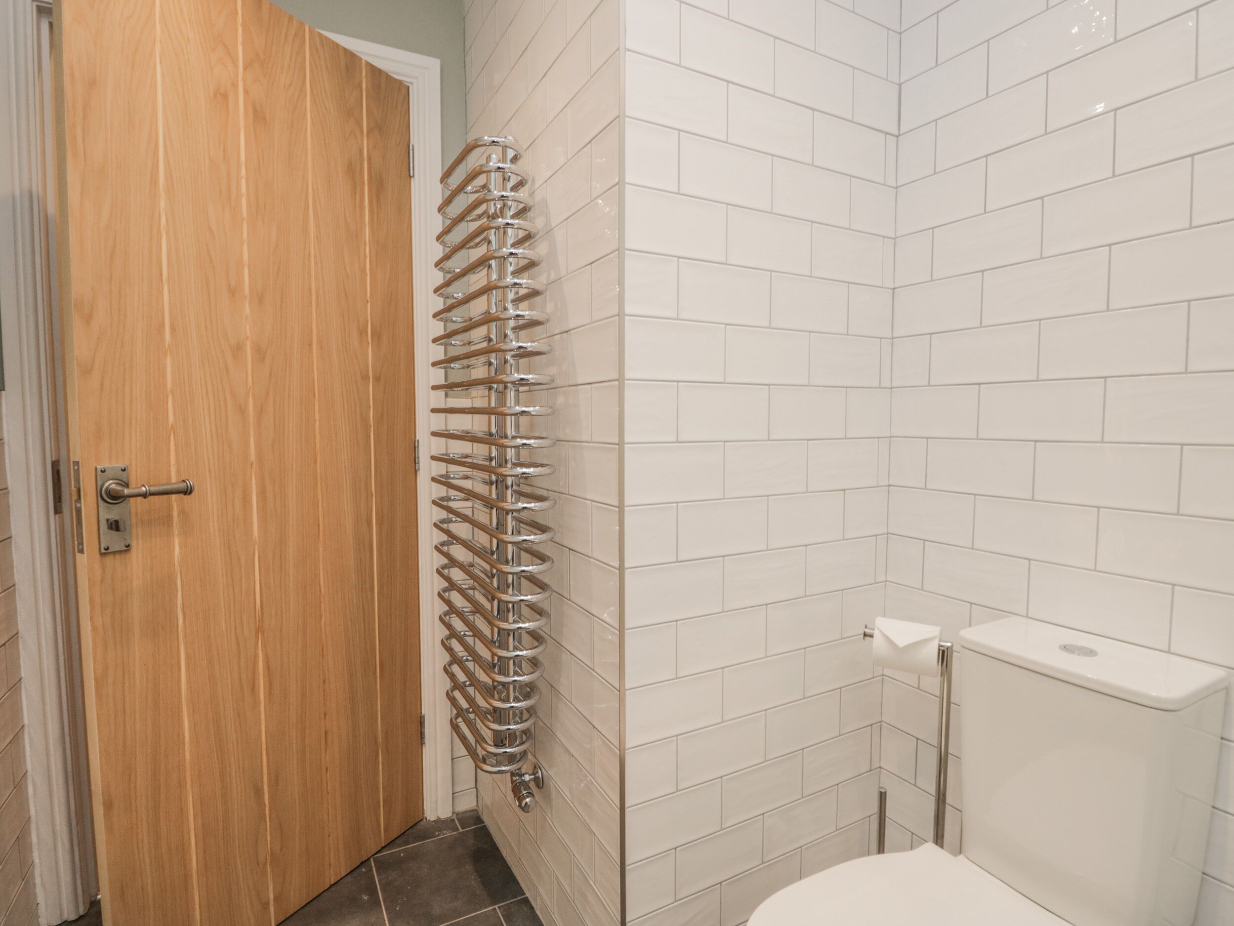 Une salle de bain avec une porte en bois, un porte-serviettes en métal, des murs recouverts de carreaux blancs et des toilettes blanches.