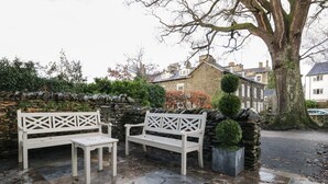 Un muro de piedra con un banco y una mesa, un árbol y una jardinera de piedra con una escultura topiaria.