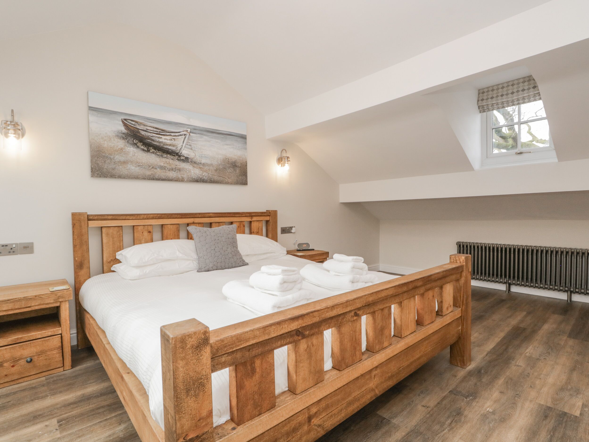 A bedroom with a wooden bed, white bedding, a painting of a boat, a skylight, and a radiator.