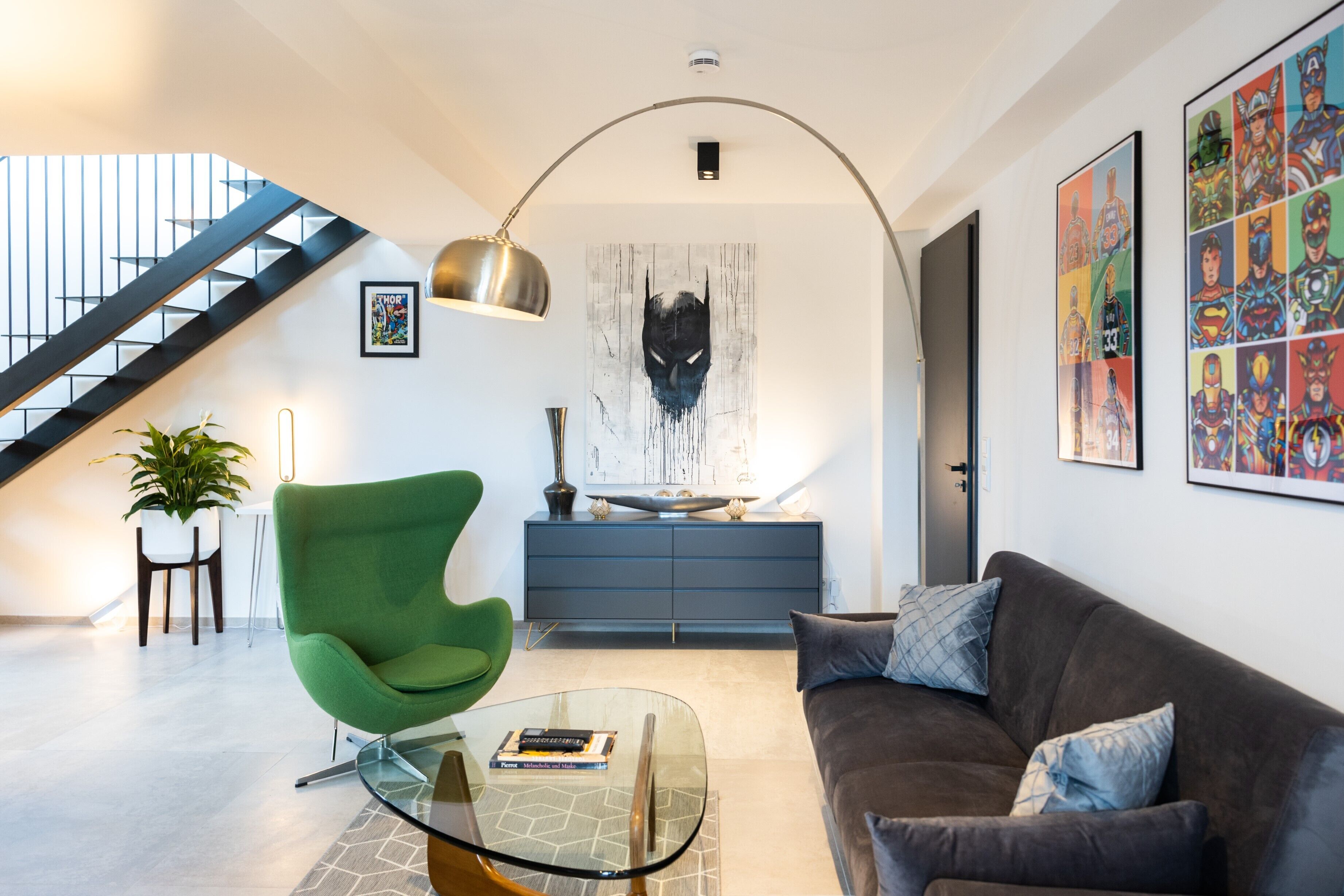 A modern living room with a green chair, a glass coffee table, a brown sofa, and a staircase with a black railing.