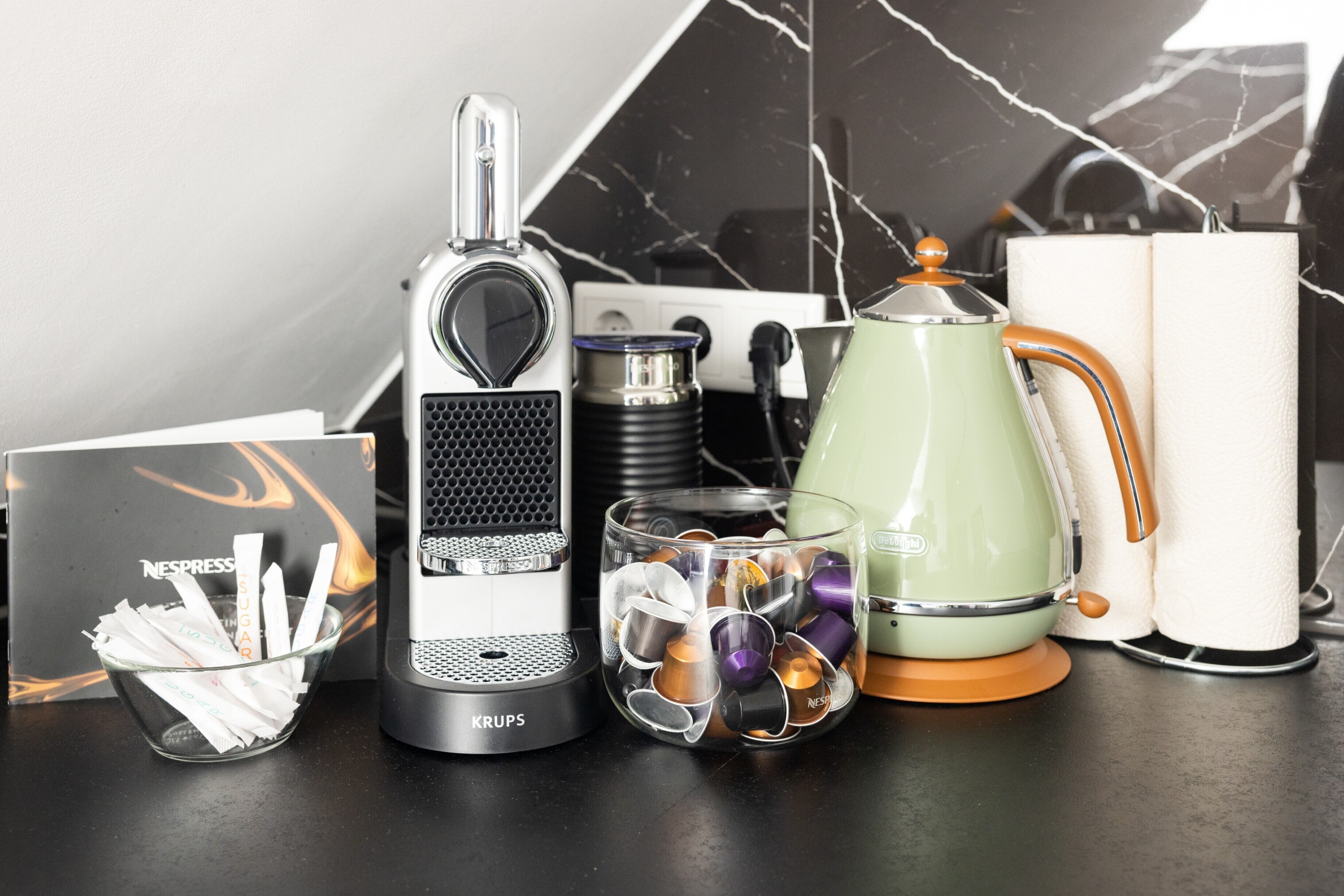 Eine Kaffeemaschine, ein Glasbehälter mit Kaffee-Kapseln, ein grüner Wasserkocher und eine Packung Nespresso-Zucker auf einer Arbeitsplatte.