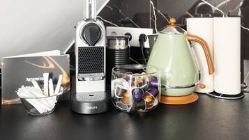 Eine Kaffeemaschine, ein Glasbehälter mit Kaffee-Kapseln, ein grüner Wasserkocher und eine Packung Nespresso-Zucker auf einer Arbeitsplatte.