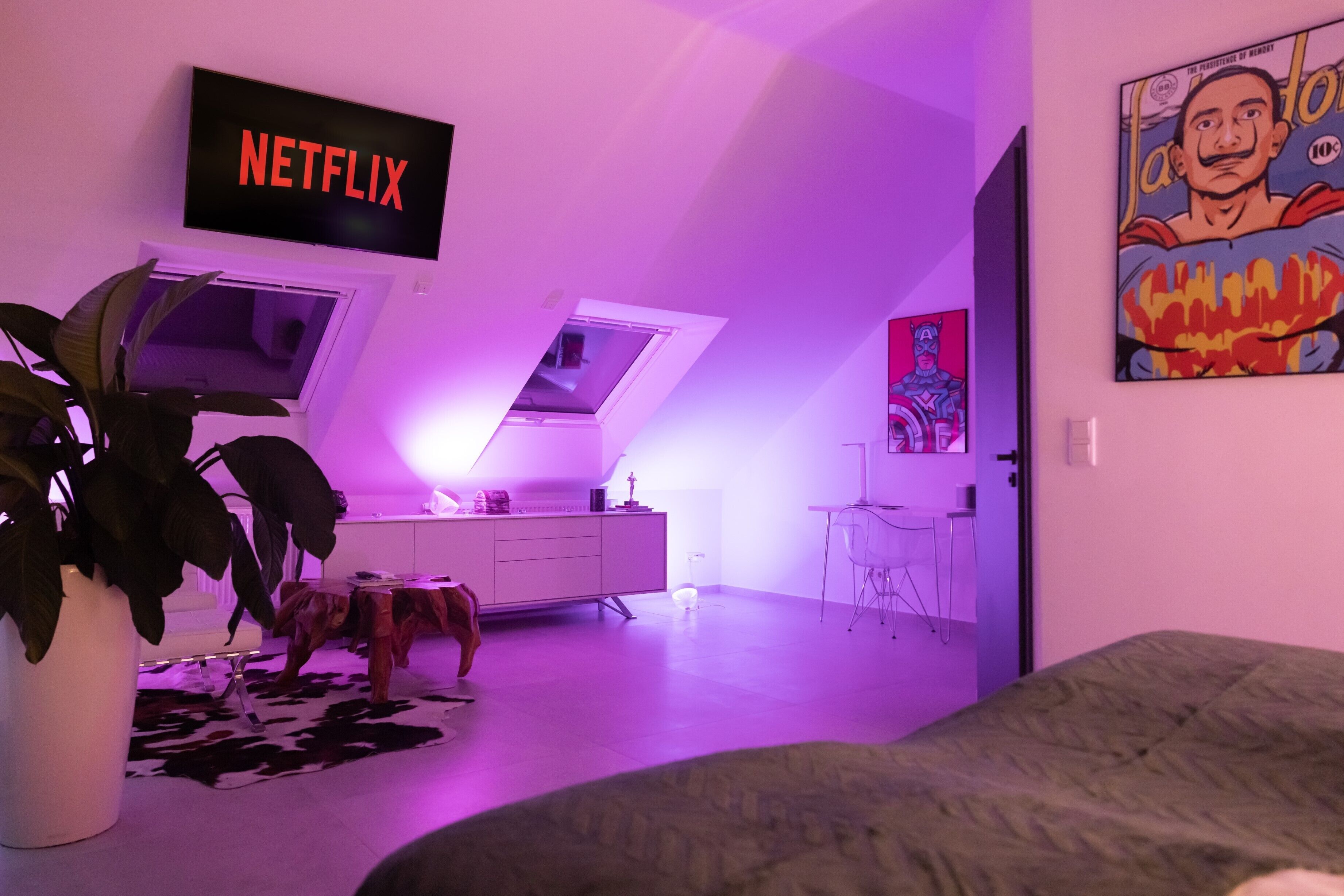 Un dormitorio moderno con techo inclinado, una planta grande en maceta, cama, escritorio y televisor mostrando 'NETFLIX'.