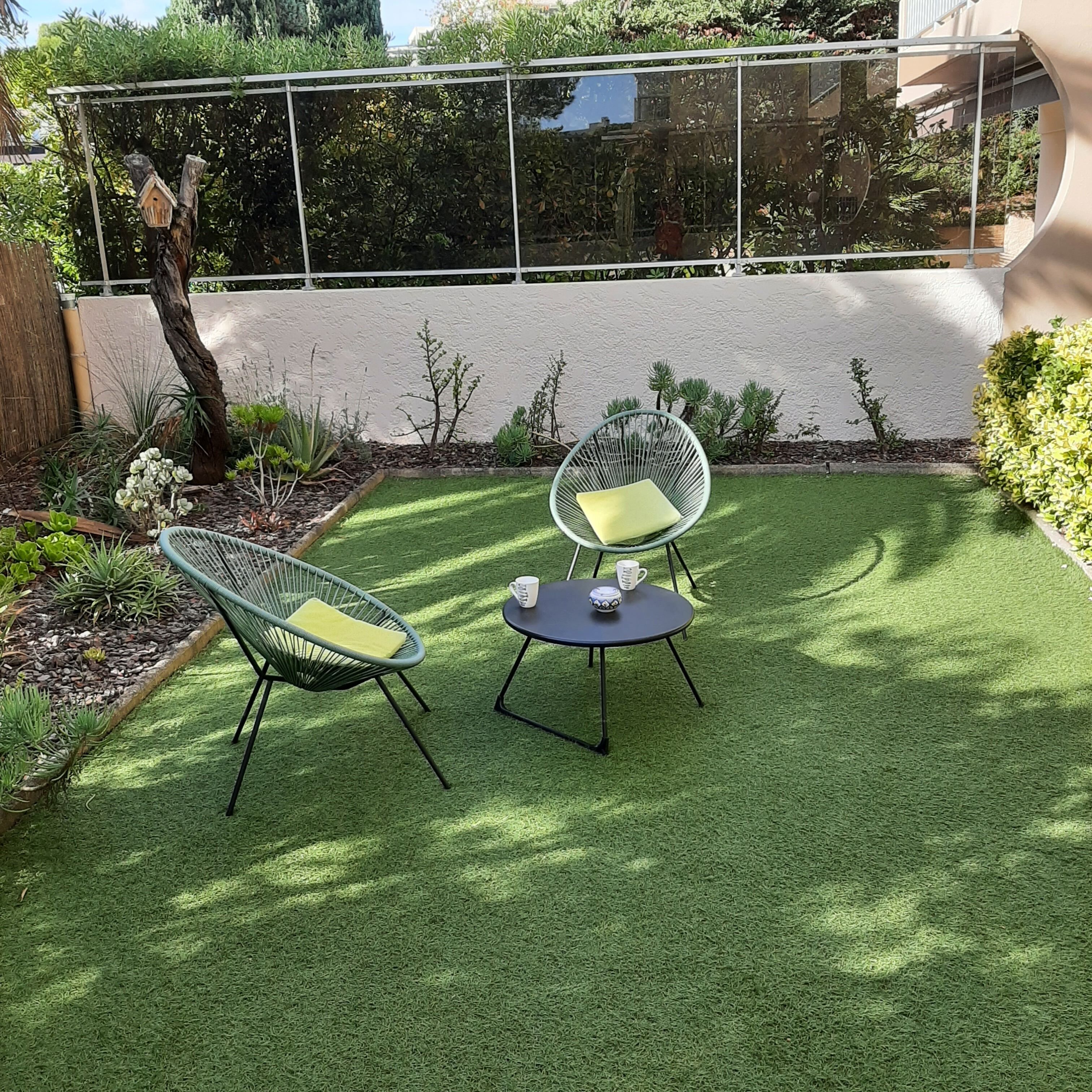 A patio with two green wire chairs and a small round table, surrounded by artificial grass and various plants.