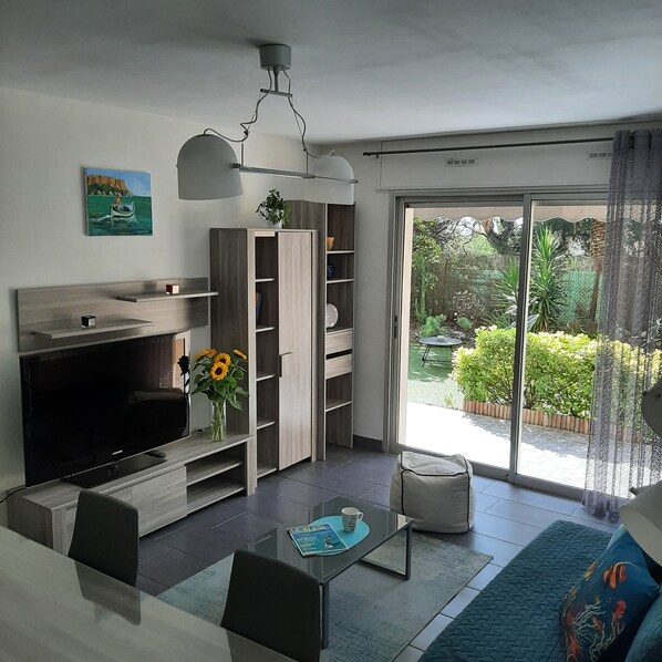 A modern living room with a flat-screen TV, a coffee table with a magazine, a sofa, and a sliding glass door leading to a garden.