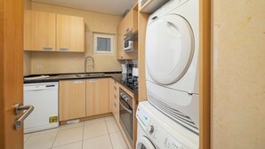 Une cuisine compacte dotée de meubles en bois, d’un four à micro-ondes encastré, d’un lave-vaisselle, ainsi que d’un lave-linge et d’un sèche-linge superposés.