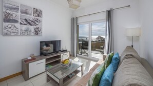 Un salon moderne doté d’un téléviseur à écran plat, d’une table basse en verre, d’un canapé avec des coussins, d’un coin repas avec des chaises et d’un balcon offrant une vue.
