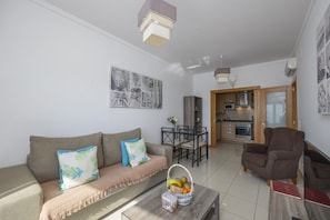 Una sala moderna con un sofá, una mesa de centro y una zona de comedor con sillas. Se ve una cocina a través de una puerta abierta.