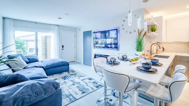 Un salon moderne avec un canapé bleu, un coin repas avec des chaises blanches et une cuisine dotée d’un plan de travail blanc.
