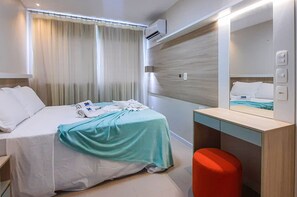 Quarto de hotel com cama, escrivaninha, espelho e um banquinho vermelho.