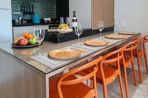 A modern kitchen with a dining area. It includes a kitchen island with a sink, a wine bottle, and a fruit bowl. There are orange chairs and a table set for dining.