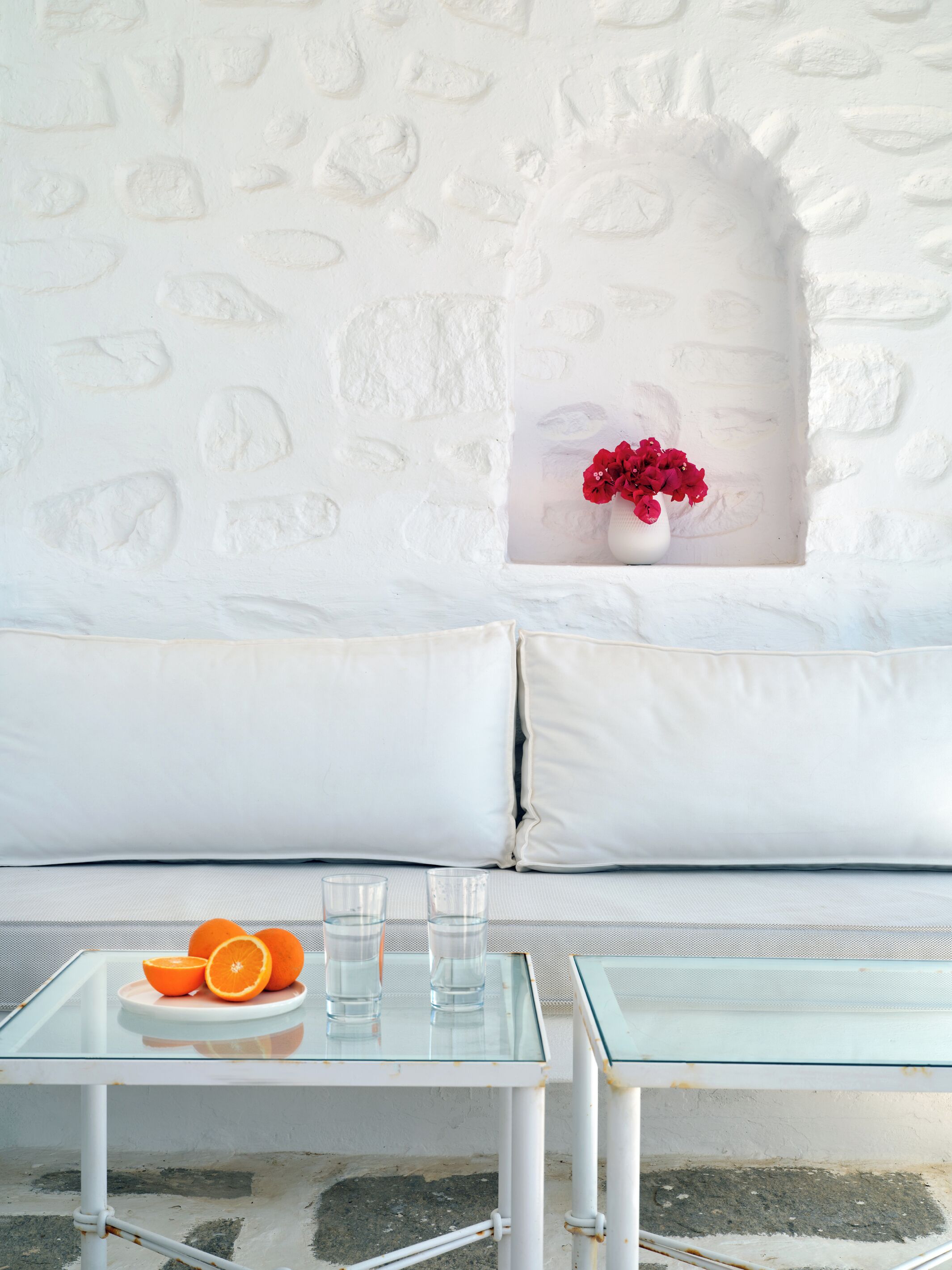 A white stone wall, a white sofa with cushions, a glass table with a bowl of oranges and glasses, and a vase with red flowers.