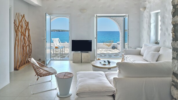 Un salón moderno con televisor, muebles blancos y vista al mar a través de amplias ventanas.