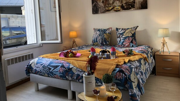 A bedroom with a bed, a window, a nightstand, a lamp, and a painting of New York City. - L'Appart 1 des Thermes (Rochefort)