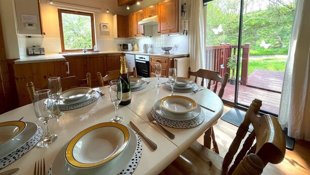 Un coin repas avec une table dressée pour quatre personnes, des meubles en bois, une cuisine en arrière-plan et une vue sur un jardin, visible à travers une porte vitrée.