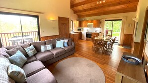 Un salon moderne avec un canapé d’angle, un coin repas avec des meubles en bois et une cuisine dotée de meubles en bois et d’une grande fenêtre.