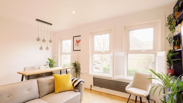A modern living room with a sofa, a dining table, and a chair. There are large windows and a radiator. - Lush Retreat with Garden - Pass the Keys (London)