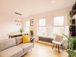 Ein modernes Wohnzimmer mit einer Couch, einem Esstisch und einem Sessel. Große Fenster und eine Heizung sind vorhanden.