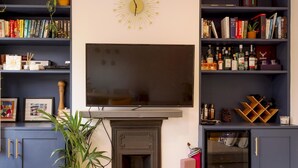 A living room with a large flat-screen TV, a fireplace, and a bookshelf filled with books and decorative items. - Lush Retreat with Garden - Pass the Keys (London)