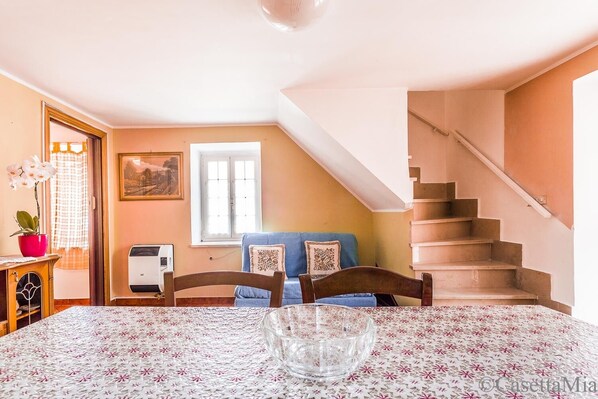 Un comedor con una mesa dispuesta para dos personas, un sofá azul, una escalera y una ventana con cortinas.
