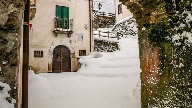 Ein schmaler Gang mit schneebedecktem Boden, ein steinernes Tor und ein Gebäude mit einer Holztür und grünen Fensterläden.