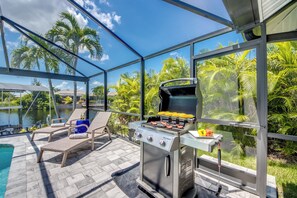 A covered outdoor area with a grill, lounge chairs, and a view of a lake and palm trees. - Villa 4Friends, modern, preferred location, heated pool, south facing (Cape Coral)