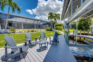 A deck with Adirondack chairs and a table, overlooking a pool and a well-maintained lawn.
