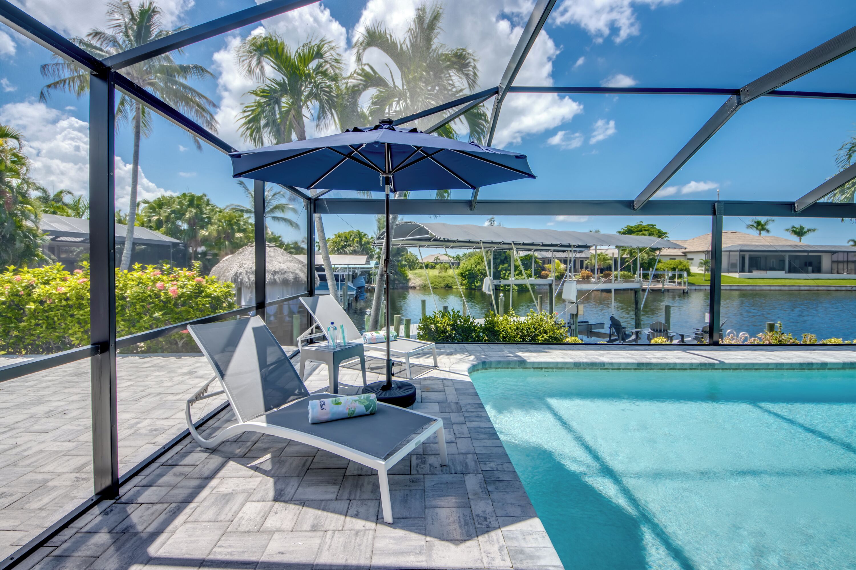 Un espace aménagé au bord de la piscine, avec des chaises longues, un coin salon couvert doté d’un parasol, et une vue sur une étendue d’eau et des palmiers.