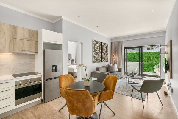 A modern kitchen with a built-in oven and refrigerator, a dining area with a round table and chairs, and a living space with a sofa and a view of a green hedge through large windows.