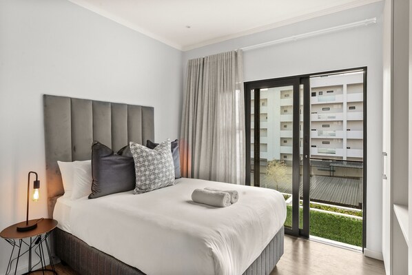 A modern hotel room with a large bed, a nightstand with a lamp, and a view of a building through the window.