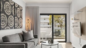 Una sala de estar moderna con un sofá gris, una mesa de centro y un televisor montado en la pared. Hay una puerta corrediza de vidrio que da a un balcón exterior con vistas a un edificio y un árbol.
