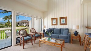 A living room with a sofa, two chairs, a coffee table, and a balcony with a view of a house and palm trees. - Manualoha 903 Steps to Poipu Beach (Koloa)
