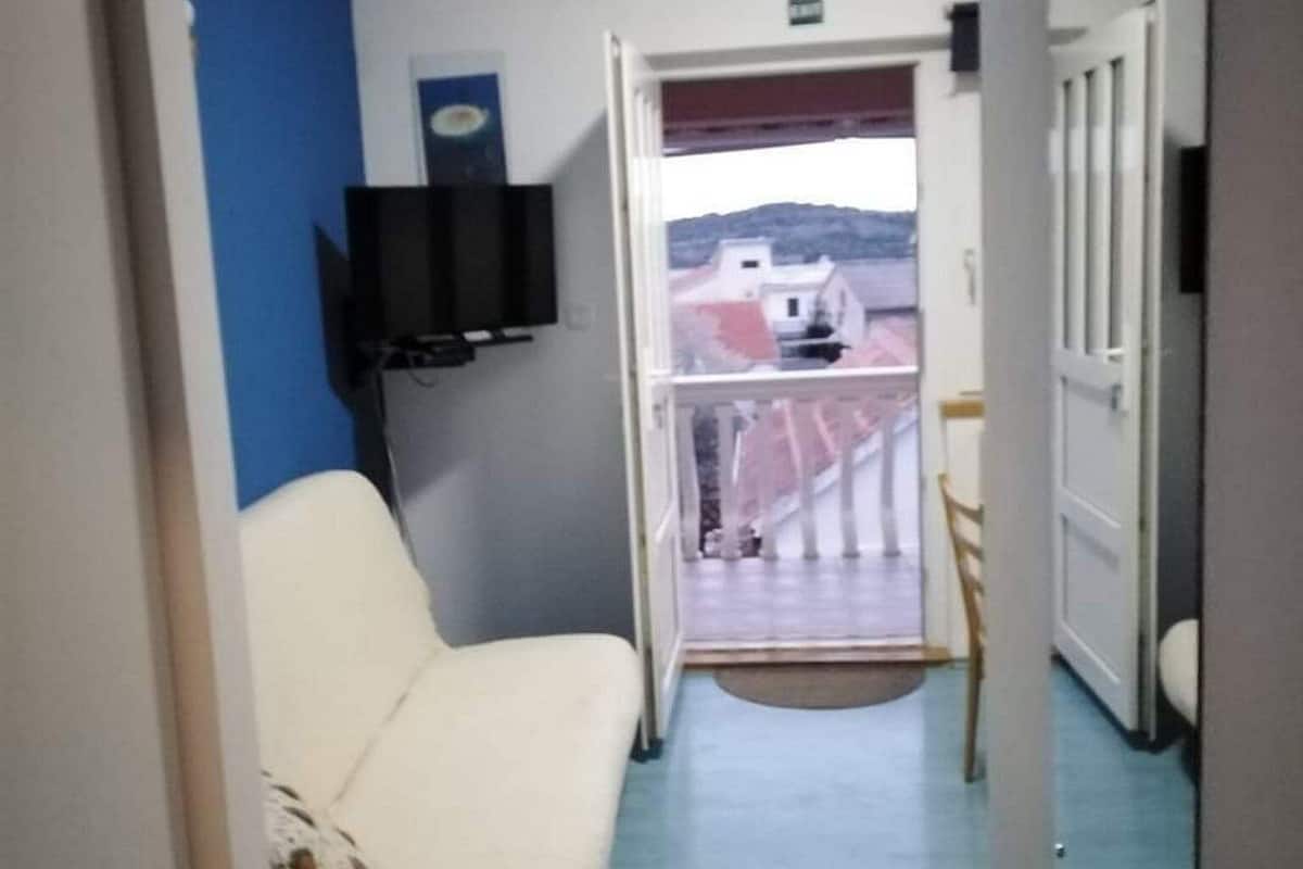Ein schmaler Flur mit einem beigen Sofa und einem Fernseher an der Wand. Eine Tür führt auf einen Balkon mit Blick auf Gebäude.