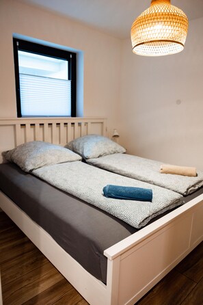 A single bed with a headboard, a window with blinds, a hanging light fixture, and a bedside table with a lamp.