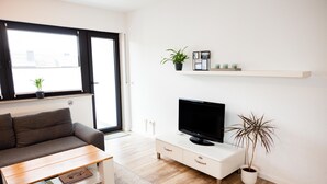 Un salon moderne comprenant un canapé, une table basse, un téléviseur à écran plat, un meuble blanc et deux plantes en pot.