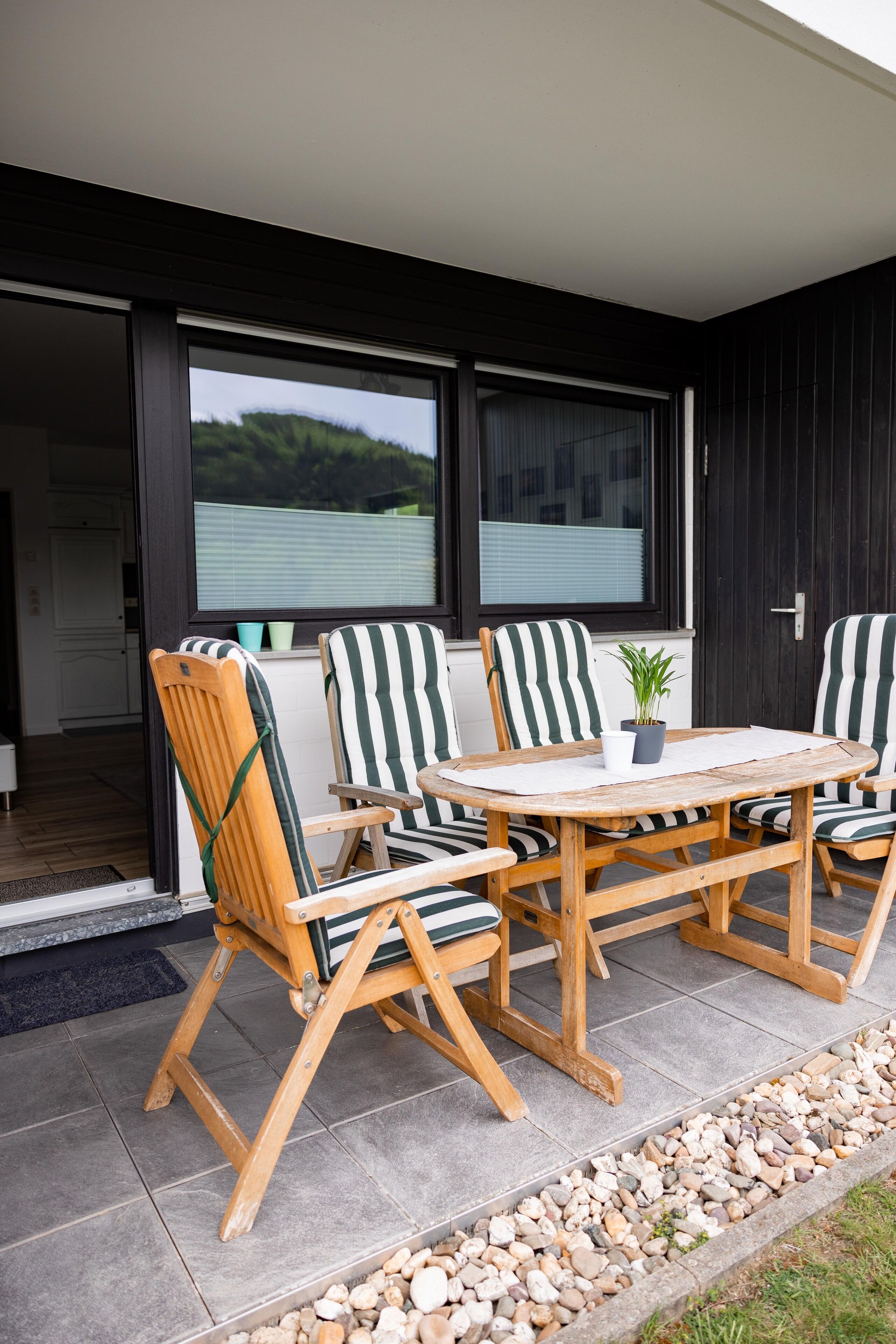 Un patio con mesa y sillas de madera, una planta en maceta y vistas a un edificio a través de grandes ventanales.