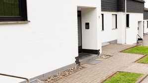 Une rangée de bâtiments modernes aux murs blancs et aux toits sombres, avec un trottoir pavé et une zone herbeuse.