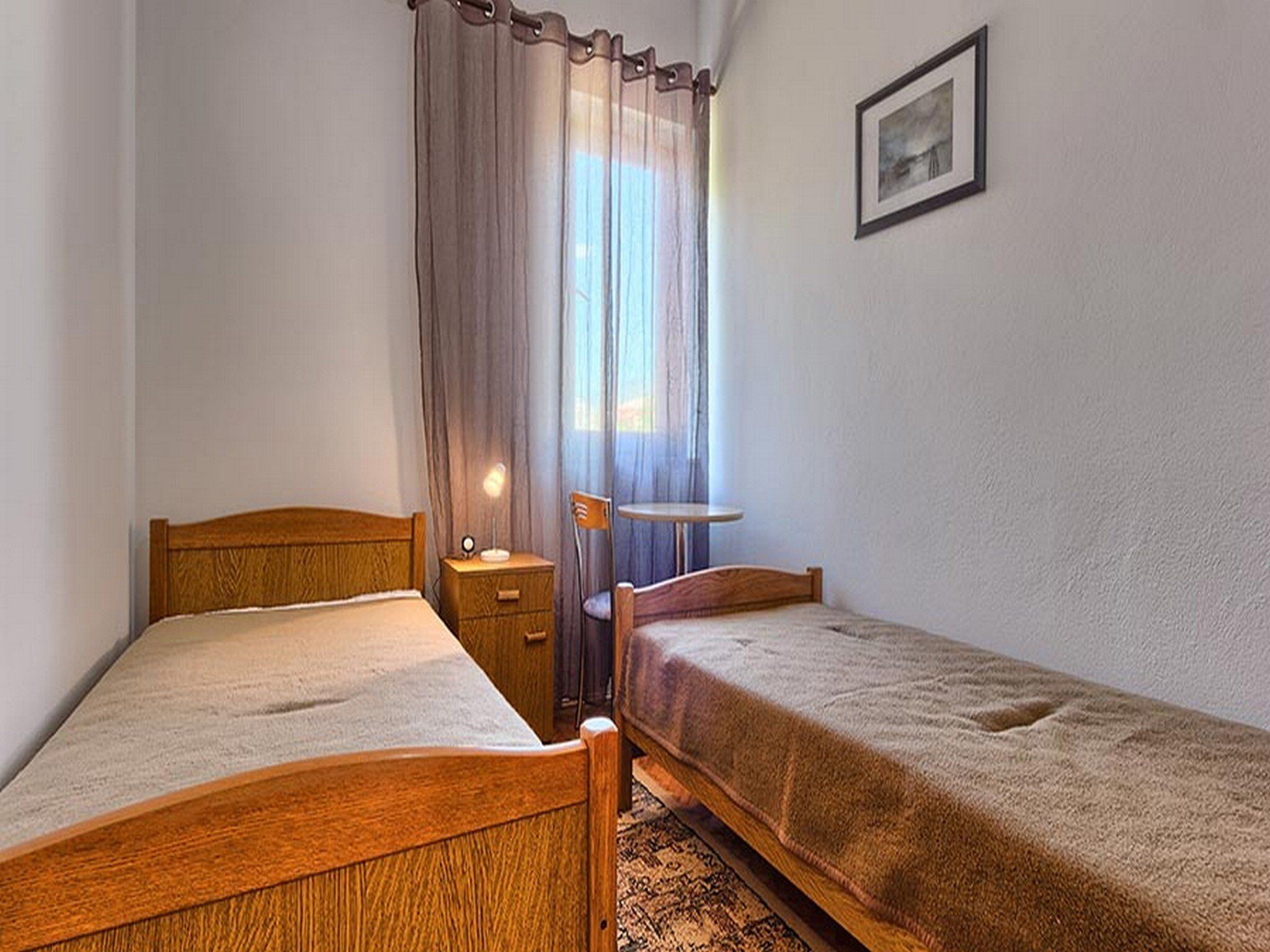 Une chambre avec deux lits en bois, une table de chevet avec une lampe, une petite table ronde et une fenêtre avec des rideaux légers.